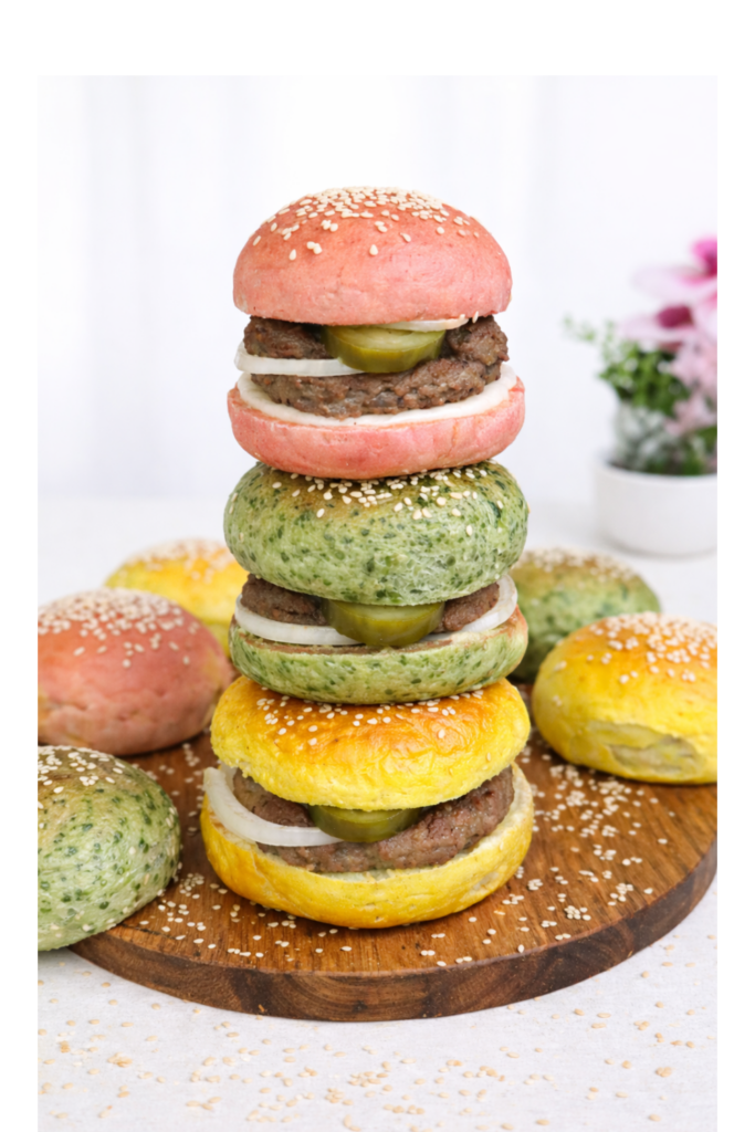 Three natural colored burger buns stacked on top of each other on a wooden board, surrounded by additional naturally colored burger buns in yellow, green, and pink tones, soft, fluffy texture, homemade bakery style presentation