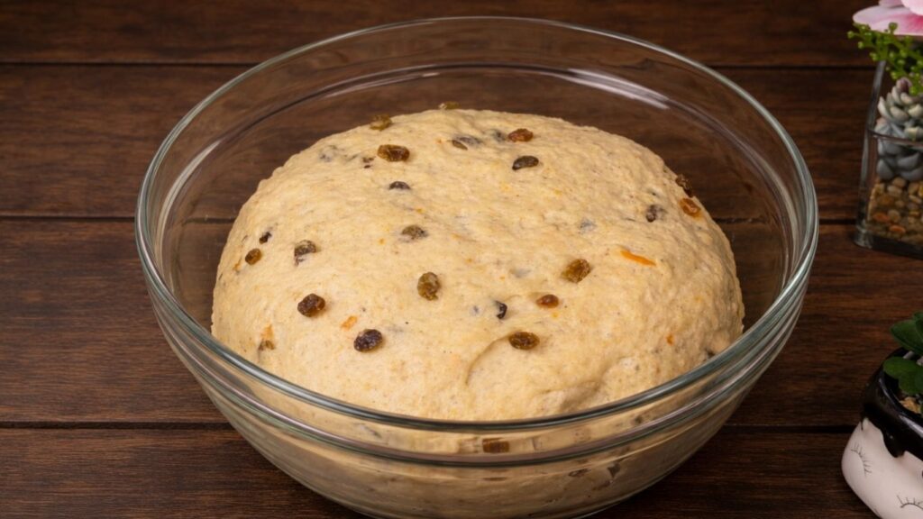 Soft and fluffy Hot Cross Buns – a smooth, soft dough resting in a clear glass bowl on a wooden table, lightly oiled and covered, ready to rise until doubled in size.