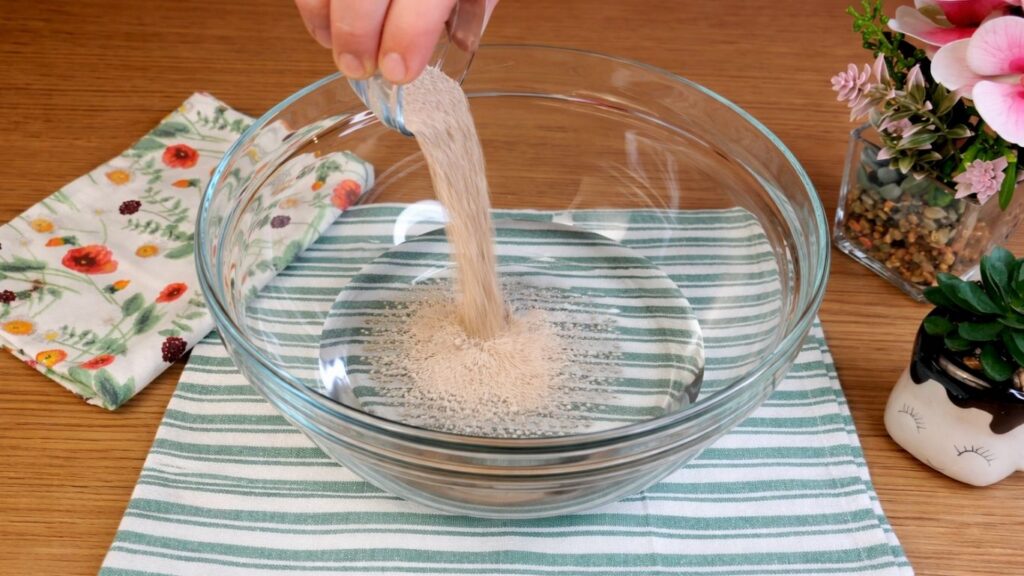 Pouring active dry yeast into a glass bowl of warm water, starting the yeast activation process for pretzel buns dough.