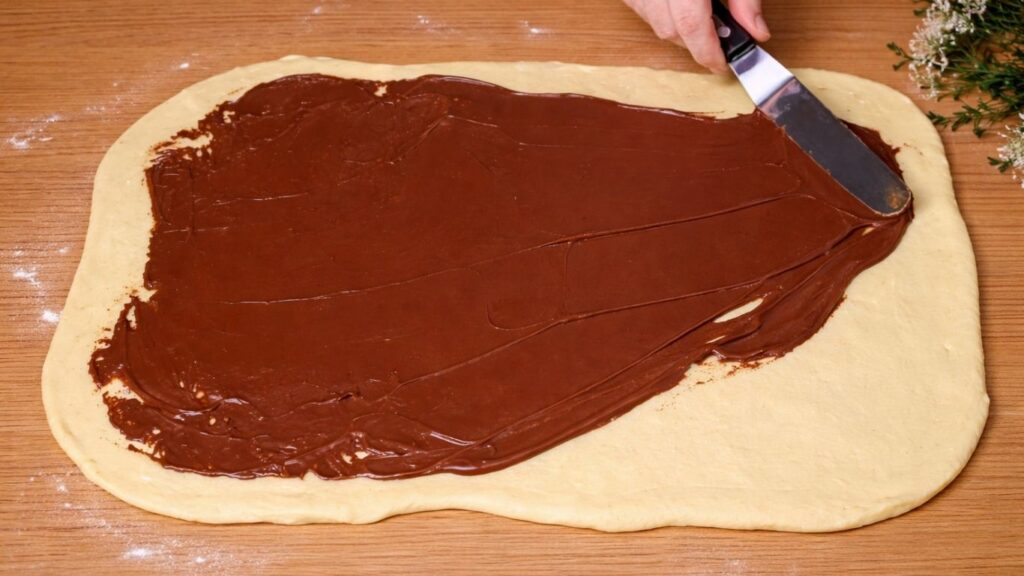 Rolled-out dough on a wooden surface being spread with chocolate filling using a spatula, evenly covering the surface for chocolate swirl buns.
