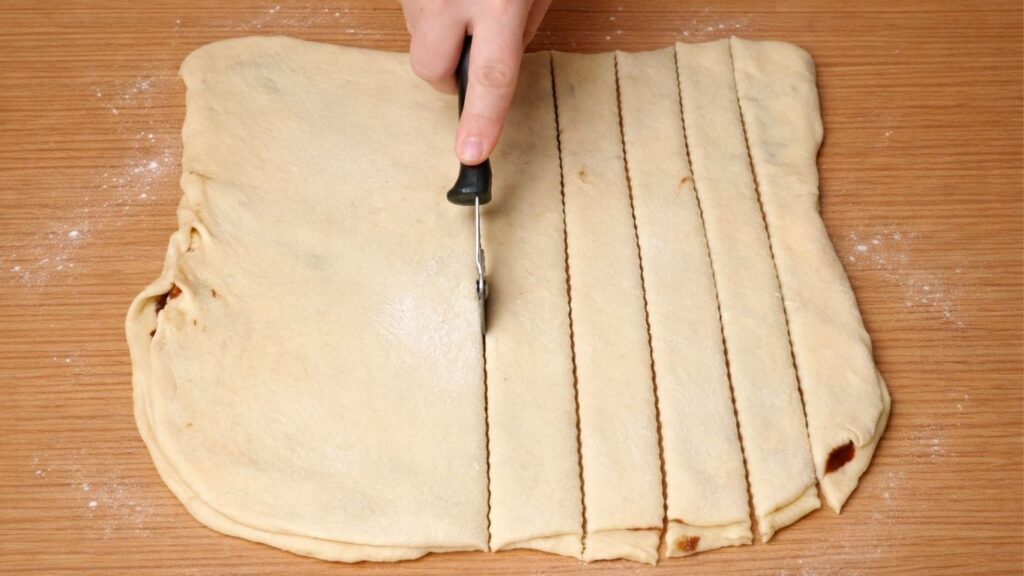 Dough folded in half and cut into strips on a wooden surface, ready to be shaped into chocolate swirl buns.