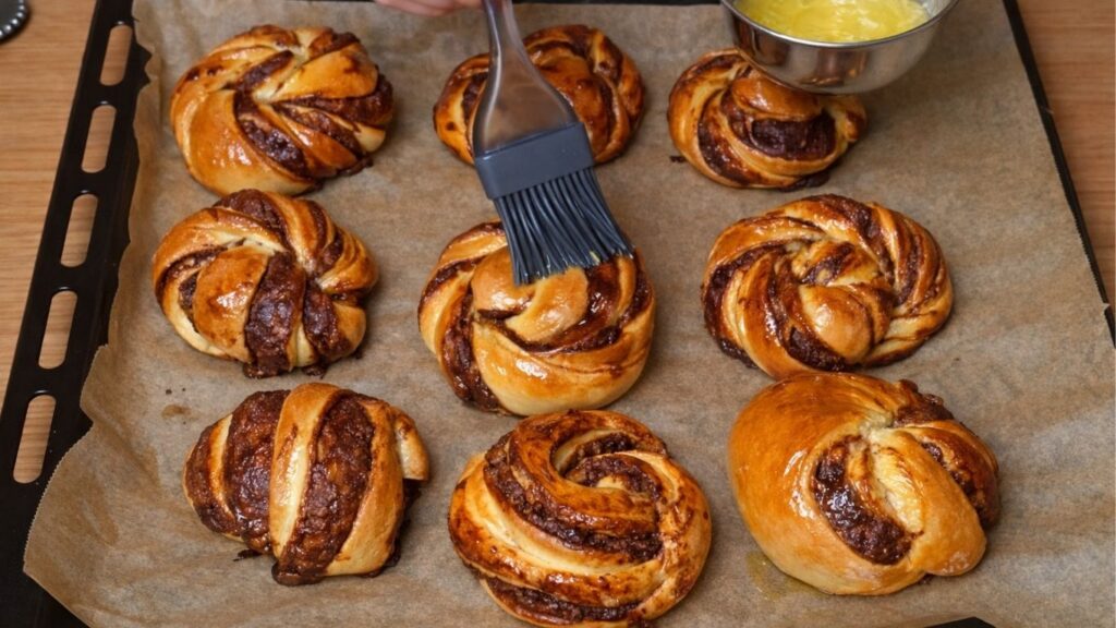 Golden-brown chocolate swirl buns being brushed with melted butter, adding shine and extra softness after baking.