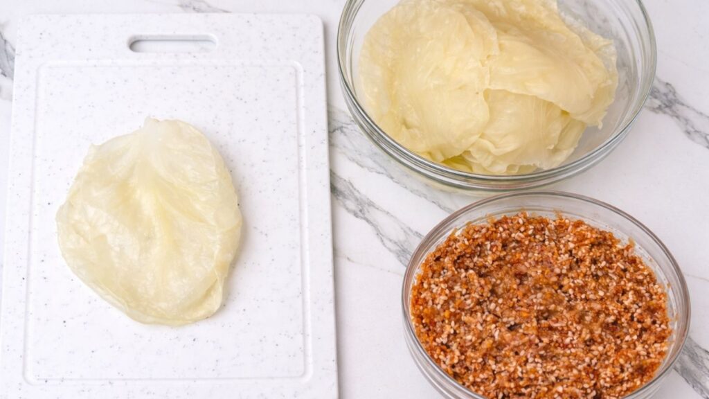 Prepared sarma ingredients on a work surface, with sour cabbage leaves in one bowl, meat and rice filling in another, and a cabbage leaf laid on a board ready for rolling stuffed cabbage rolls