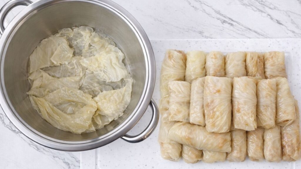 Pot lined with cabbage leaves on the bottom, with rolled sarma on a board beside it, ready to be arranged and cooked as traditional stuffed sour cabbage rolls