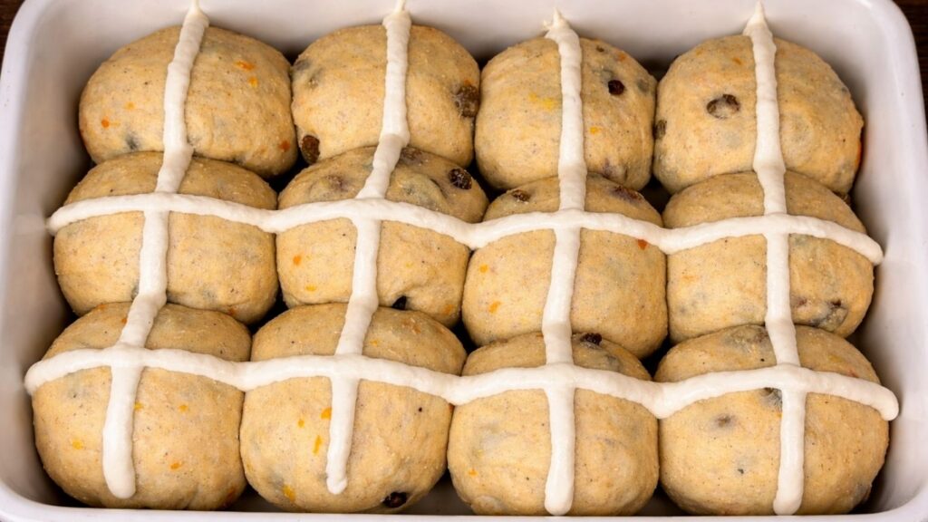 Soft and fluffy Hot Cross Buns – shaped dough balls arranged on a baking tray with white cross patterns piped on top of each bun, ready to bake until golden and fragrant.