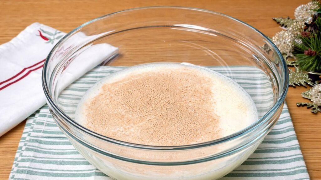 Glass bowl with milk, sugar, and yeast mixed together, showing activated yeast that has risen and become foamy on the surface.