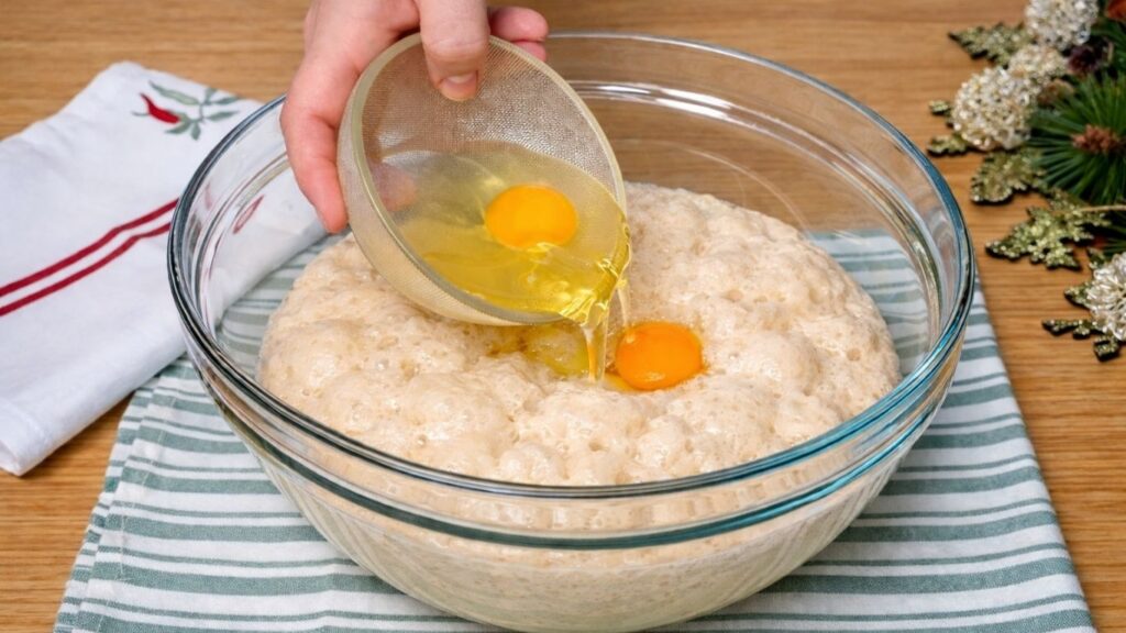 Glass bowl with activated, risen yeast as eggs are being poured into the mixture, ready for the next step of the dough preparation.