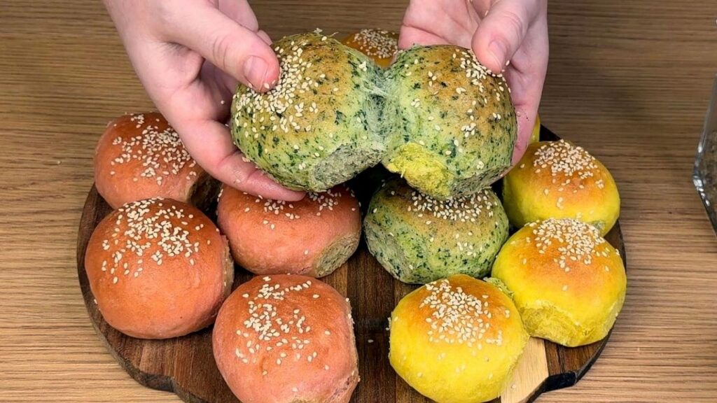 All baked burger buns arranged on a wooden board as hands pull apart two buns to reveal the soft, fluffy interior, colorful yellow, green, and pink tones, clean and natural homemade baking scene