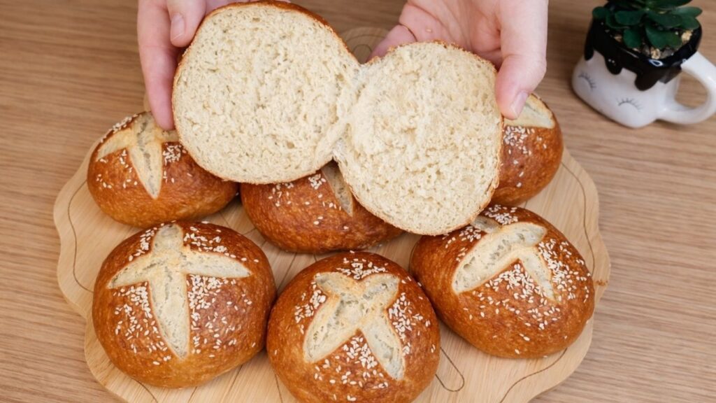 Freshly baked pretzel buns stacked on a wooden board, with one bun cut in half to show the soft, fluffy interior and golden pretzel crust.