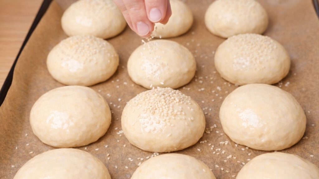Sprinkling sesame seeds over the glazed buns on a parchment-lined baking tray, just before placing them in the oven to bake until golden and fluffy