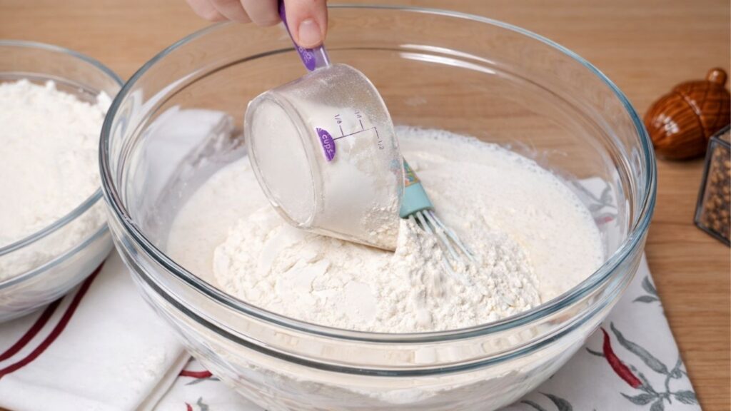 Glass bowl with activated yeast mixture (milk and sugar) as flour is being poured in, showing the start of mixing the dough for soft and fluffy burger buns