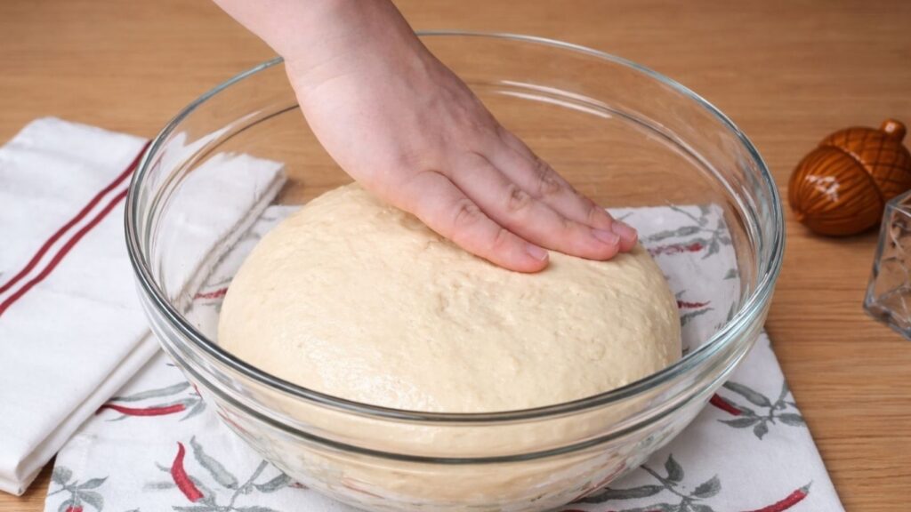 Smooth dough placed in a lightly oiled glass bowl, with a hand gently brushing oil over the top to keep it soft and prevent drying, ready to rest covered for 1 hour until doubled in size