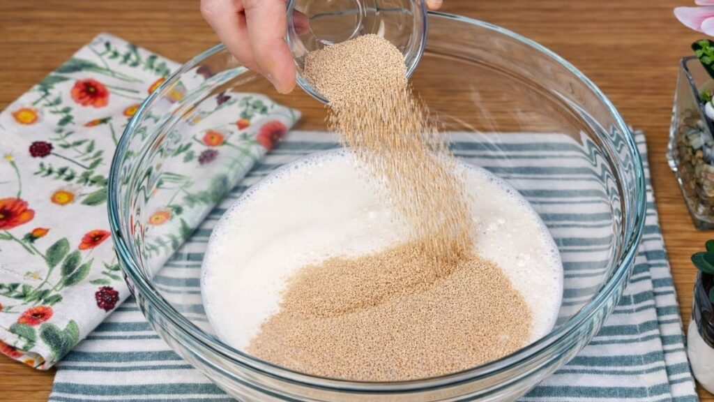 Glass bowl with warm milk and sugar as active dry yeast is being poured in, starting the yeast activation process, close-up, clean and natural baking preparation scene