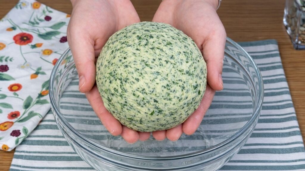 Hands holding soft green dough above a glass bowl, naturally colored with blended spinach, smooth and elastic texture, clean and natural baking preparation scene