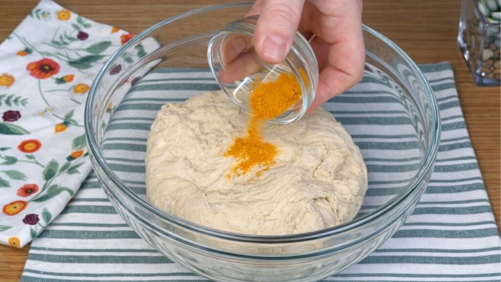Glass bowl with one portion of dough as turmeric is being added to color it naturally yellow, close-up clean baking preparation scene