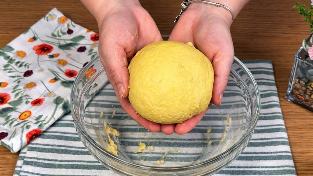 Hands holding soft yellow dough above a glass bowl, naturally colored with turmeric, smooth and elastic texture, clean and natural baking preparation scene
