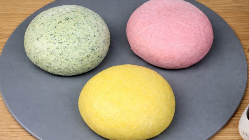 Three portions of kneaded dough in yellow, green, and pink on a silicone mat, shaped and ready to rest and rise for one hour, clean and natural baking preparation scene