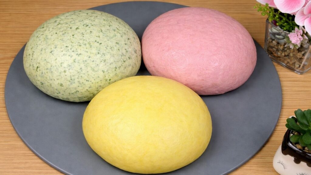 Three portions of naturally colored dough—yellow, green, and pink—fully risen on a silicone mat, soft, airy, and doubled in size, ready for shaping, clean and natural baking scene