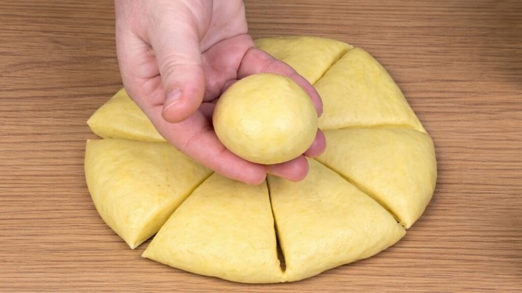 Yellow dough on a clean work surface divided into seven portions while the eighth piece is shaped into a smooth dough ball held in hand, ready for burger buns, natural baking preparation scene