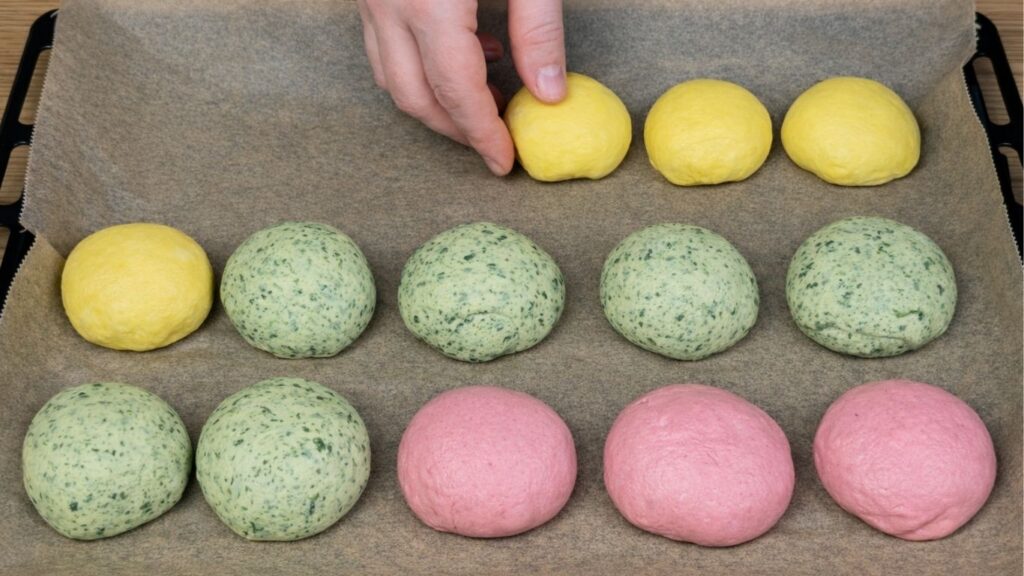 Naturally colored burger buns arranged in a baking tray lined with parchment paper, yellow, green, and pink buns neatly placed and ready for proofing, clean and natural baking preparation scene