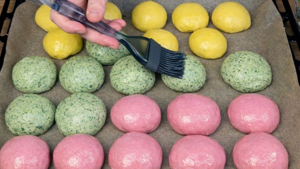 Naturally colored burger buns arranged in a baking tray lined with parchment paper as a brush applies a mixture of water and oil on top, preparing them for baking, clean and natural baking preparation scene