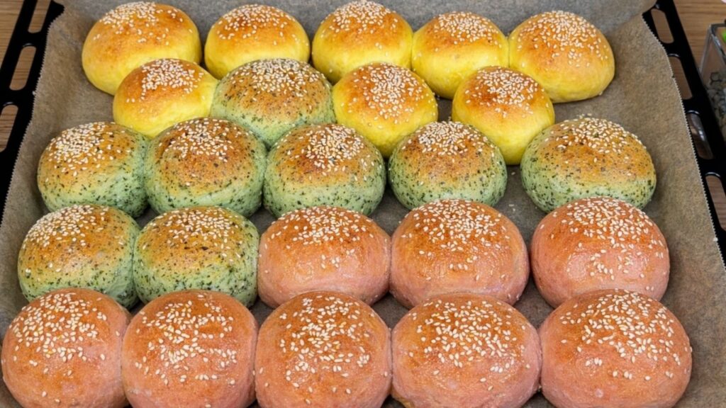 Freshly baked naturally colored burger buns in a baking tray lined with parchment paper, beautifully golden on top with vibrant yellow, green, and pink tones, soft and fluffy homemade bakery style scene
