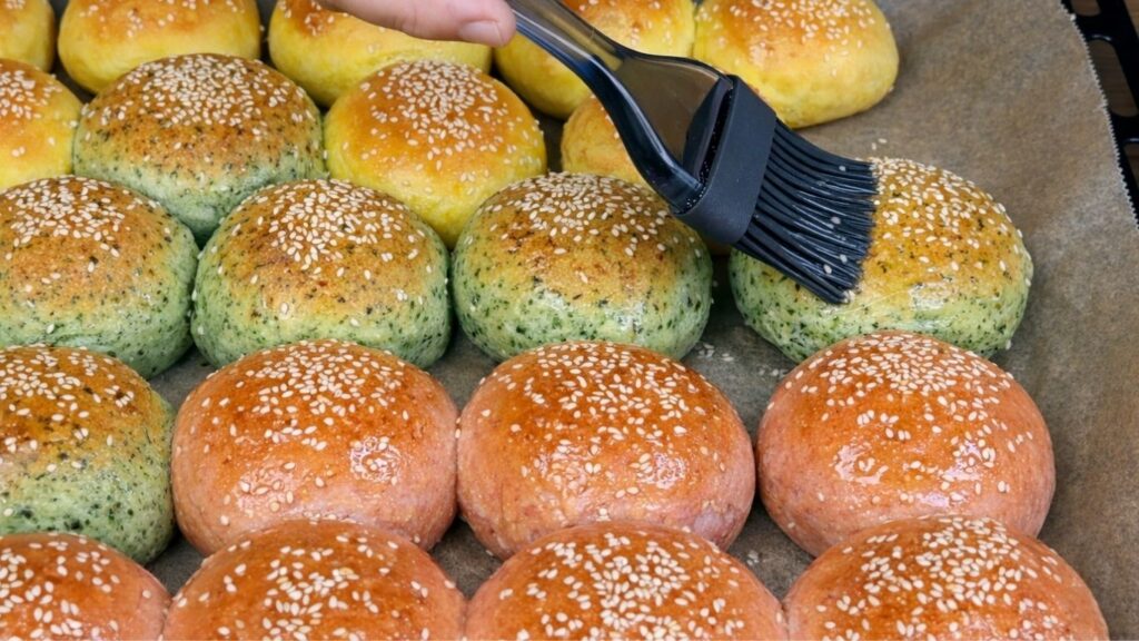 Freshly baked colorful burger buns in a baking tray lined with parchment paper as a silicone brush applies a water and oil glaze on top, giving them a soft shine, clean and natural homemade baking scene