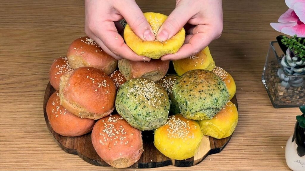 All burger buns arranged on a wooden board as hands gently press a yellow bun to show how soft and fluffy it is, naturally colored yellow, green, and pink tones, clean and natural homemade baking scene