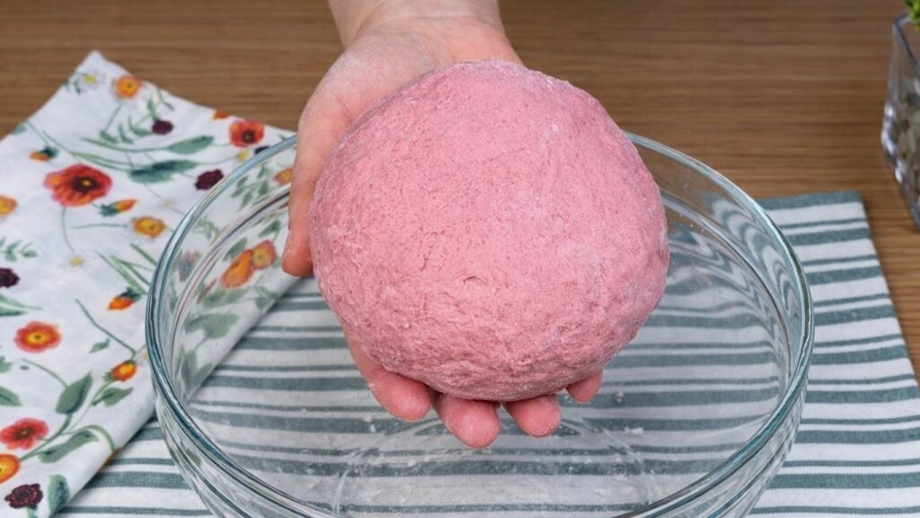 Hands holding soft pink dough above a glass bowl, naturally colored with blended beetroot, smooth and elastic texture, clean and natural baking preparation scene