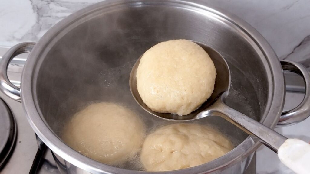 Removing boiled pretzel buns from the pot with a slotted spoon after baking soda bath.
