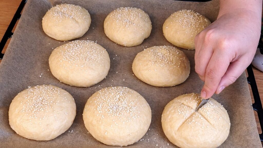 Pretzel buns arranged on a baking tray, sprinkled with sesame seeds and scored with cross cuts before baking.