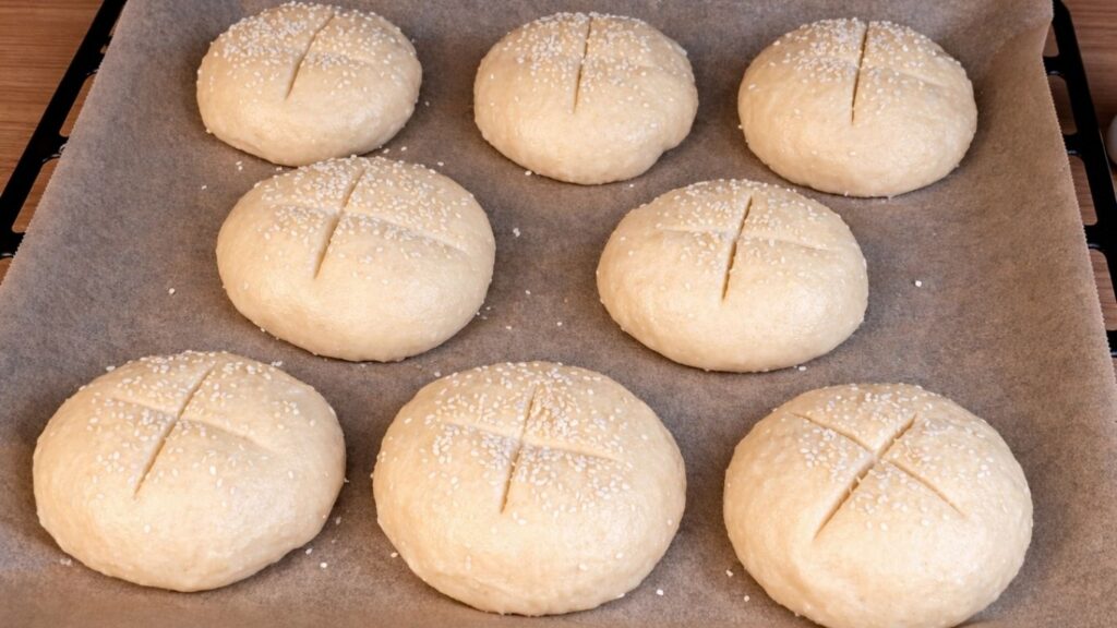 Boiled pretzel buns on parchment paper, topped with sesame seeds and scored with cross cuts, ready for baking.