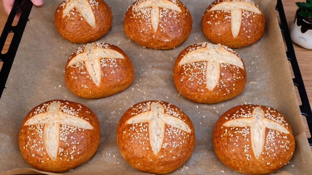 Freshly baked pretzel buns on parchment paper in a baking tray, golden brown with a classic pretzel crust.