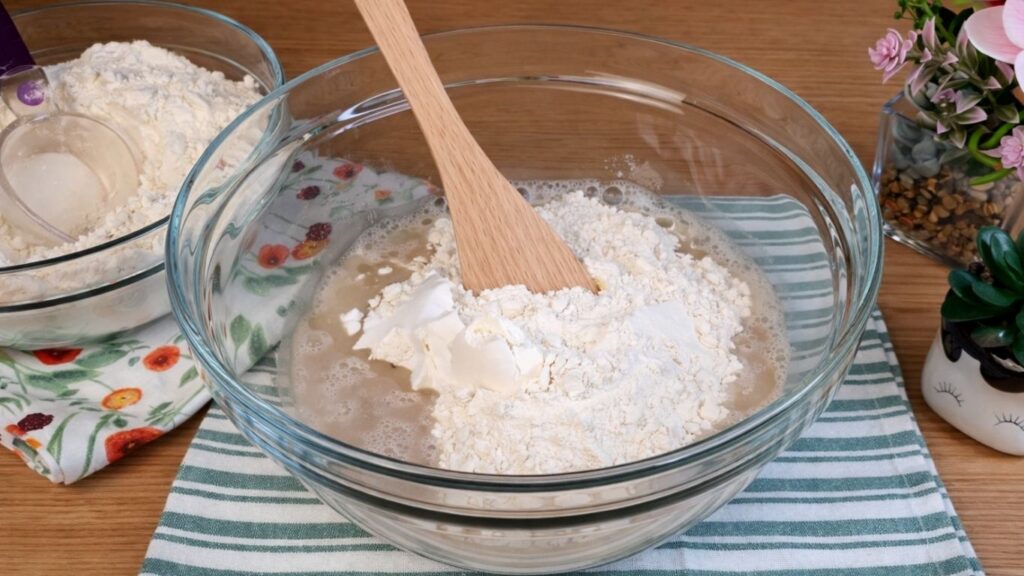 Adding flour, oil and salt to the activated yeast mixture in a glass bowl to start forming pretzel buns dough.