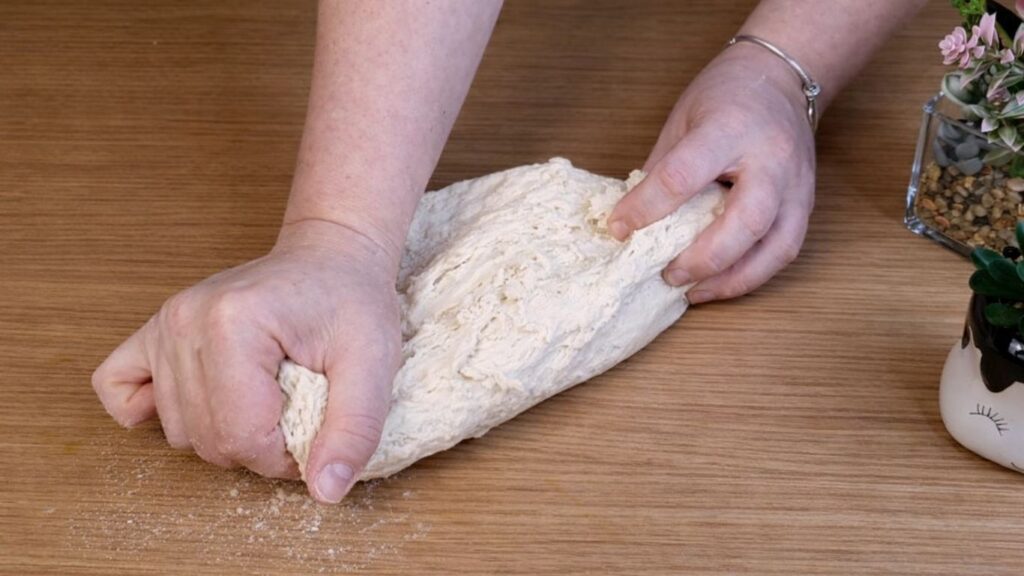 Kneading and stretching pretzel buns dough by hand on a wooden work surface until smooth and elastic.