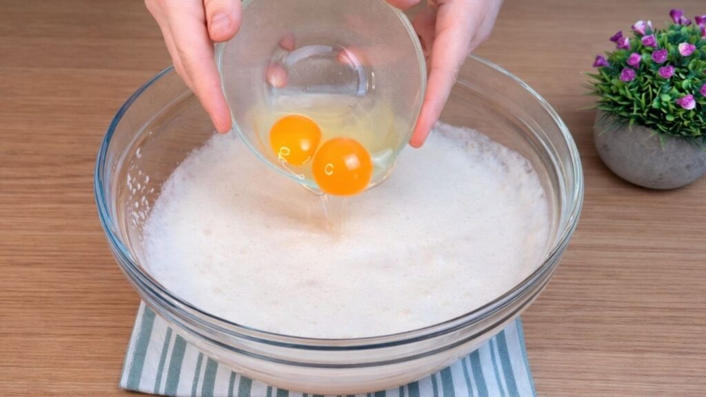 Adding two eggs to the activated yeast mixture for homemade hot dog buns dough.