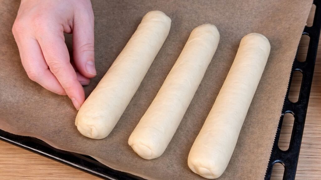 Placing shaped homemade hot dog buns onto a lined baking tray before baking.