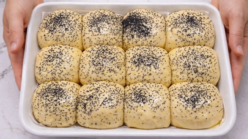 brushed dinner rolls topped with poppy seeds in a baking tray ready for baking