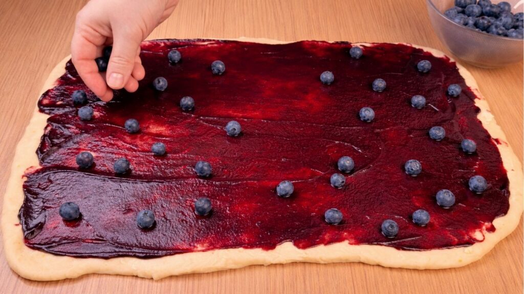 sprinkling fresh blueberries over the blueberry jam on rolled dough for blueberry lemon rolls