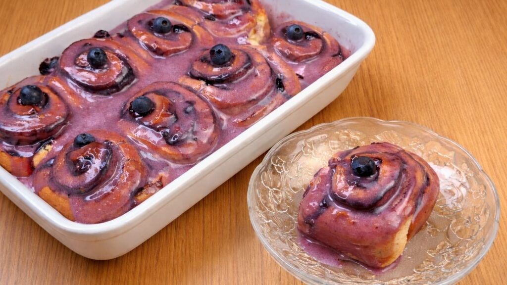 blueberry lemon rolls covered with cream cheese frosting in a baking pan with one roll served on a plate