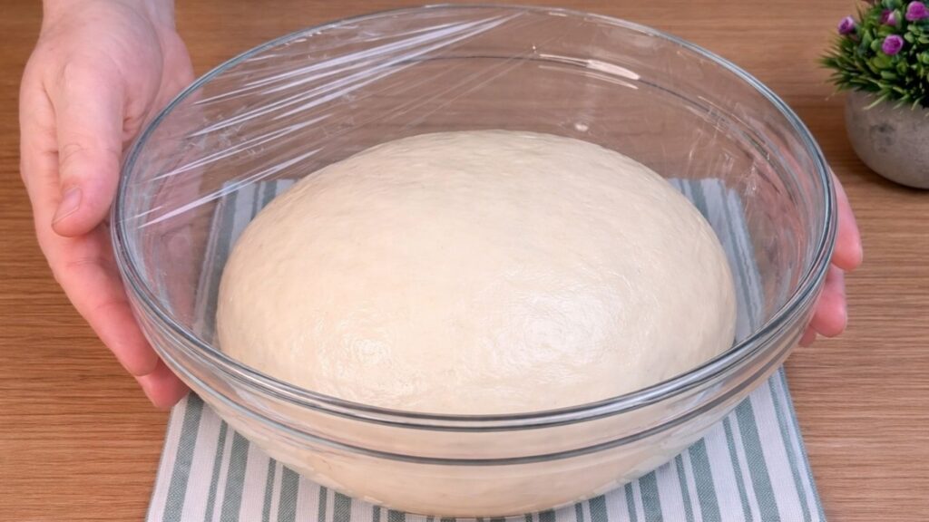 Dough for homemade hot dog buns covered with plastic wrap and left to rise in a bowl.