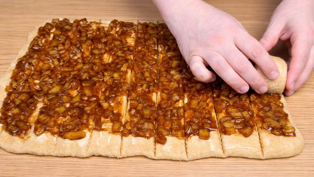 Rolling strips of dough with apple filling to shape homemade apple cinnamon rolls.