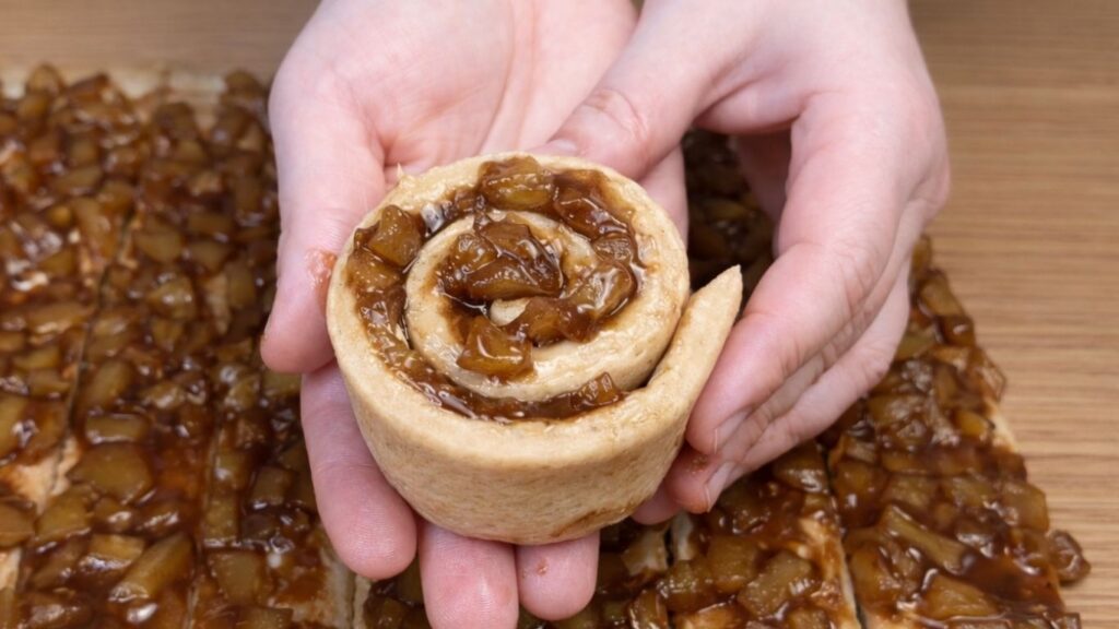 Hand holding a freshly shaped apple cinnamon roll before placing it in the baking dish.