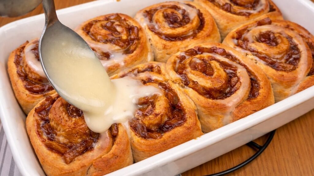 Spreading cream cheese frosting over warm apple cinnamon rolls in the baking dish.