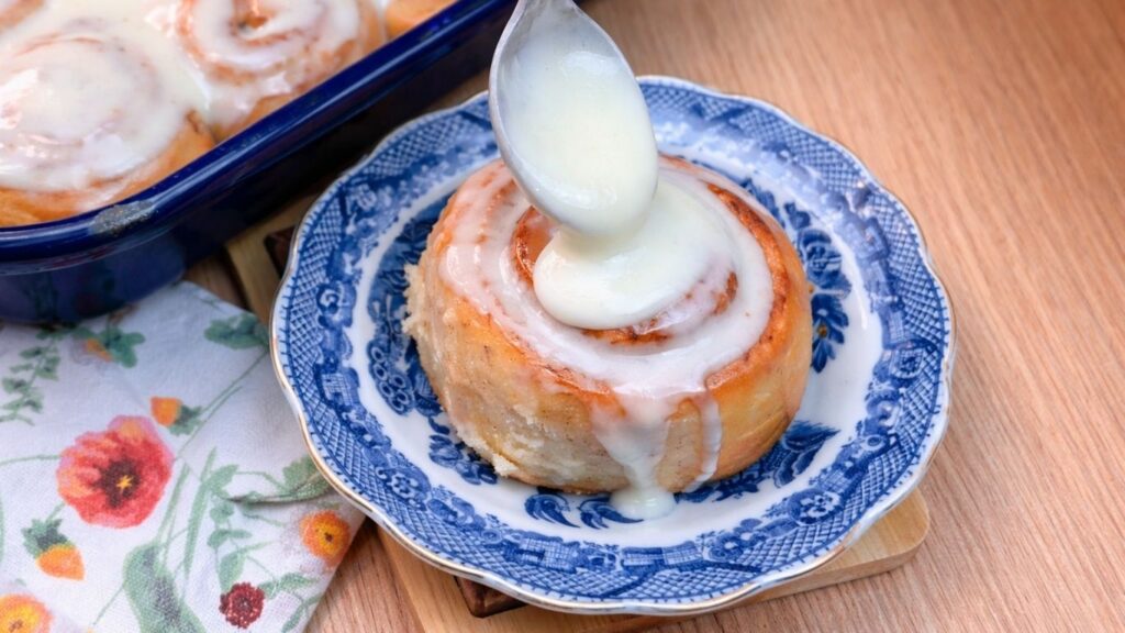 pouring cream cheese glaze over a homemade cinnamon roll served on a plate