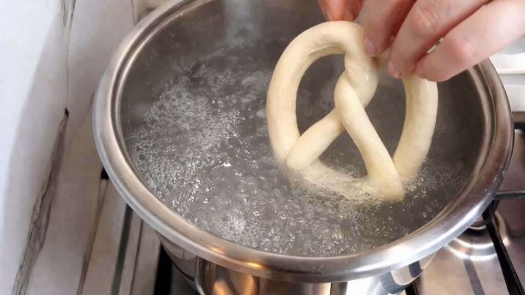 Carefully placing a homemade pretzel into the boiling baking soda water bath.