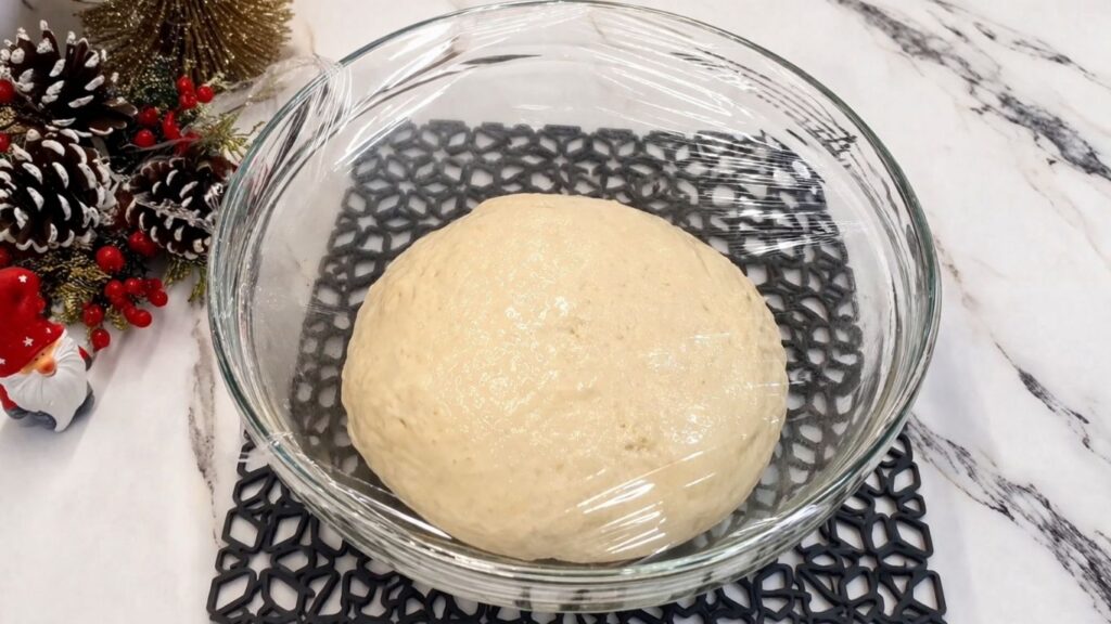 Homemade pretzel dough covered with plastic wrap resting and rising in a greased bowl.