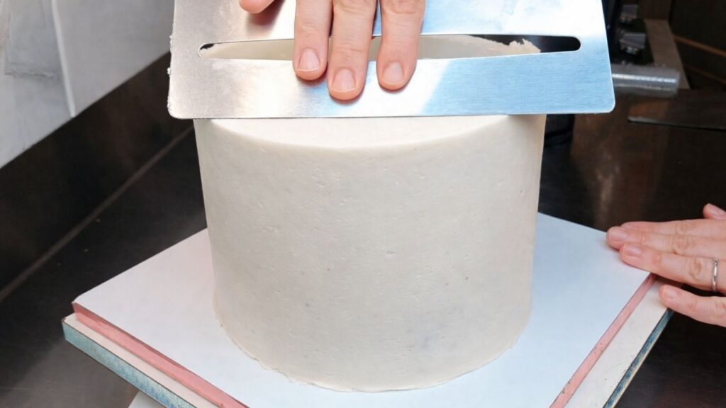 Smoothing the buttercream with a scraper to level the cake and prepare it for candy cake decorating.