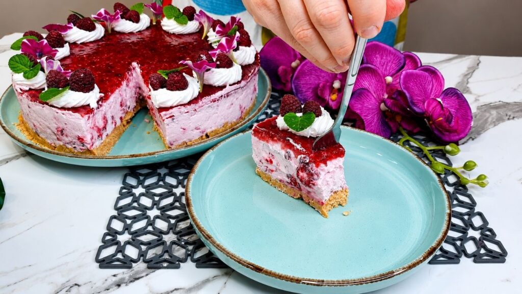 No bake raspberry yogurt cake recipe sliced on turquoise plate showing
creamy yogurt layers, graham cracker crust, and fresh raspberries with
whipped cream topping