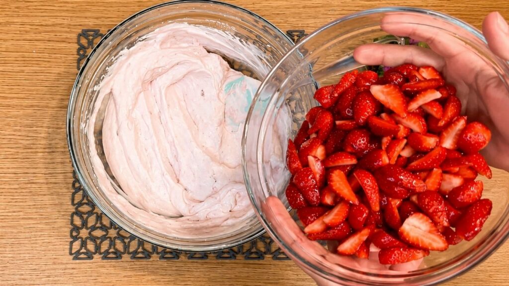 Adding strawberry yogurt, sugar, vanilla extract, and chopped strawberries into the whipped cream, then mixing gently until the filling is smooth and evenly combined.